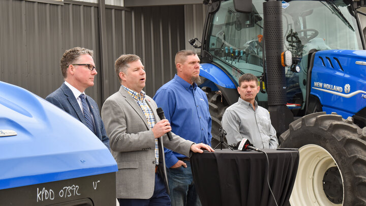 A man speaks into a microphone as he stands next to three other men between two New Holland methane-powered tractors.