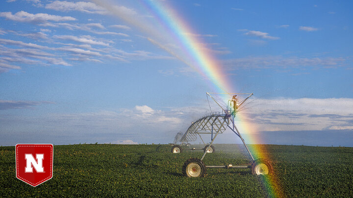 A rainbow forms over an irrigation pivot in a grain field, with a blue sky in the background.